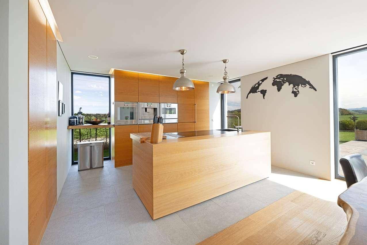 Moderne Holzküche mit Kochinsel, integrierten Geräten und viel Tageslicht durch große Fenster.