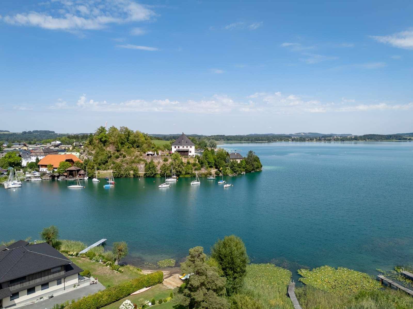 Weitläufiger Blick auf einen ruhigen See mit Segelbooten, einer malerischen Uferlandschaft und Häusern unter blauem Himmel.