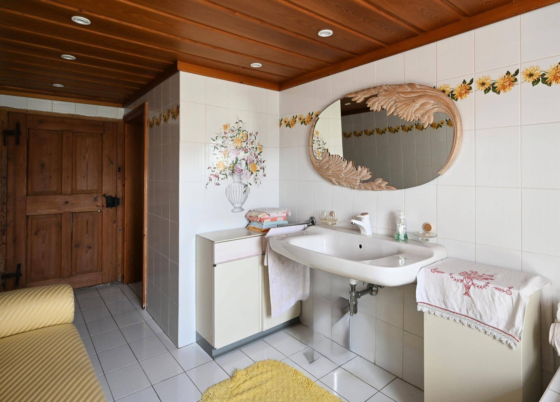 Helles Badezimmer mit weißen Fliesen, Holzdecke und einem dekorativen Spiegel.