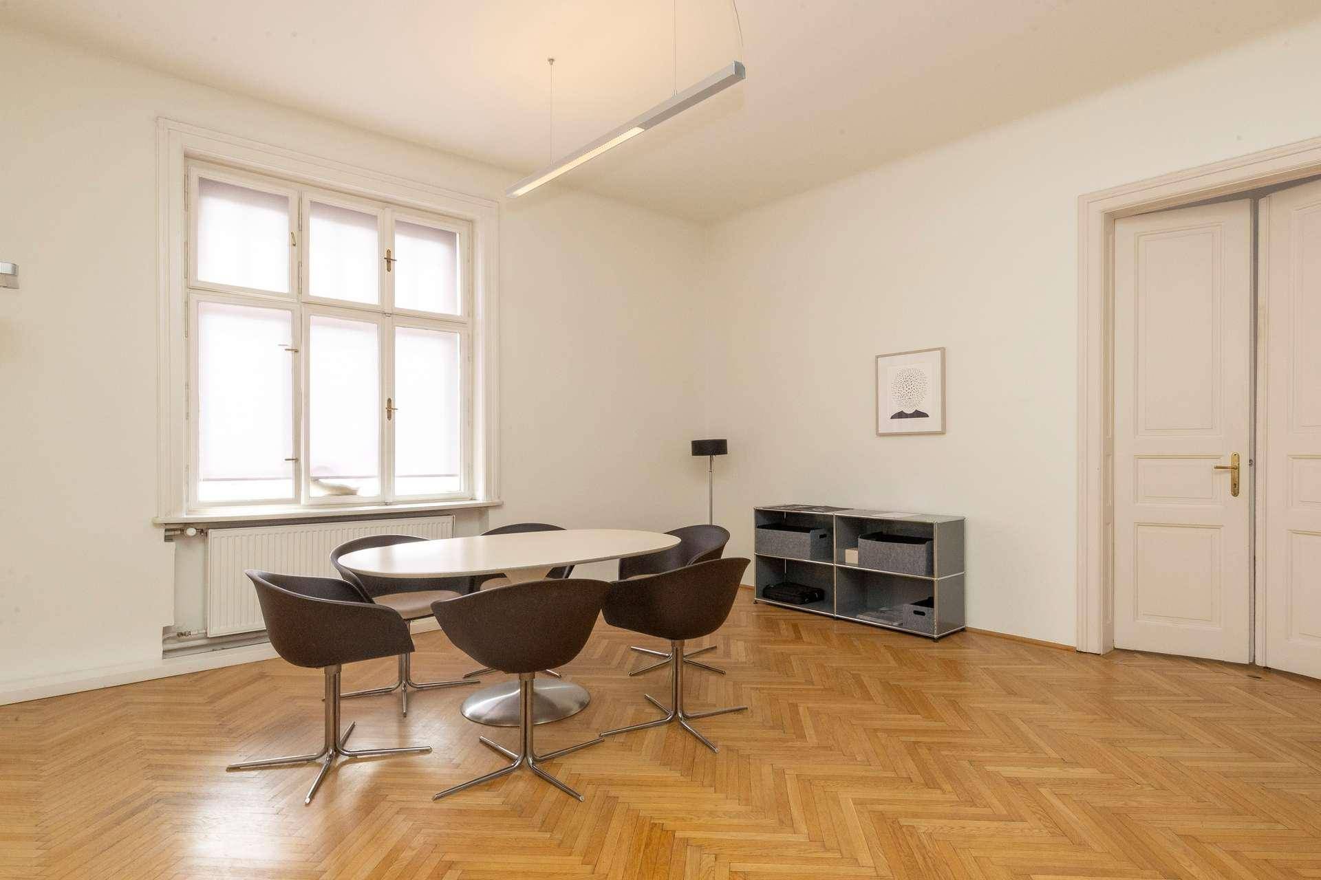 Helles Büro mit ovalem Tisch, modernen Stühlen, Fischgrätparkett und großem Fenster mit Jalousien.