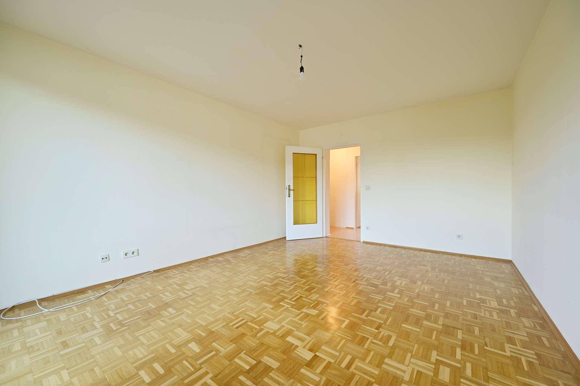Geräumiges, leeres Zimmer mit klassischem Parkettboden und einer Tür mit Glaseinsatz.