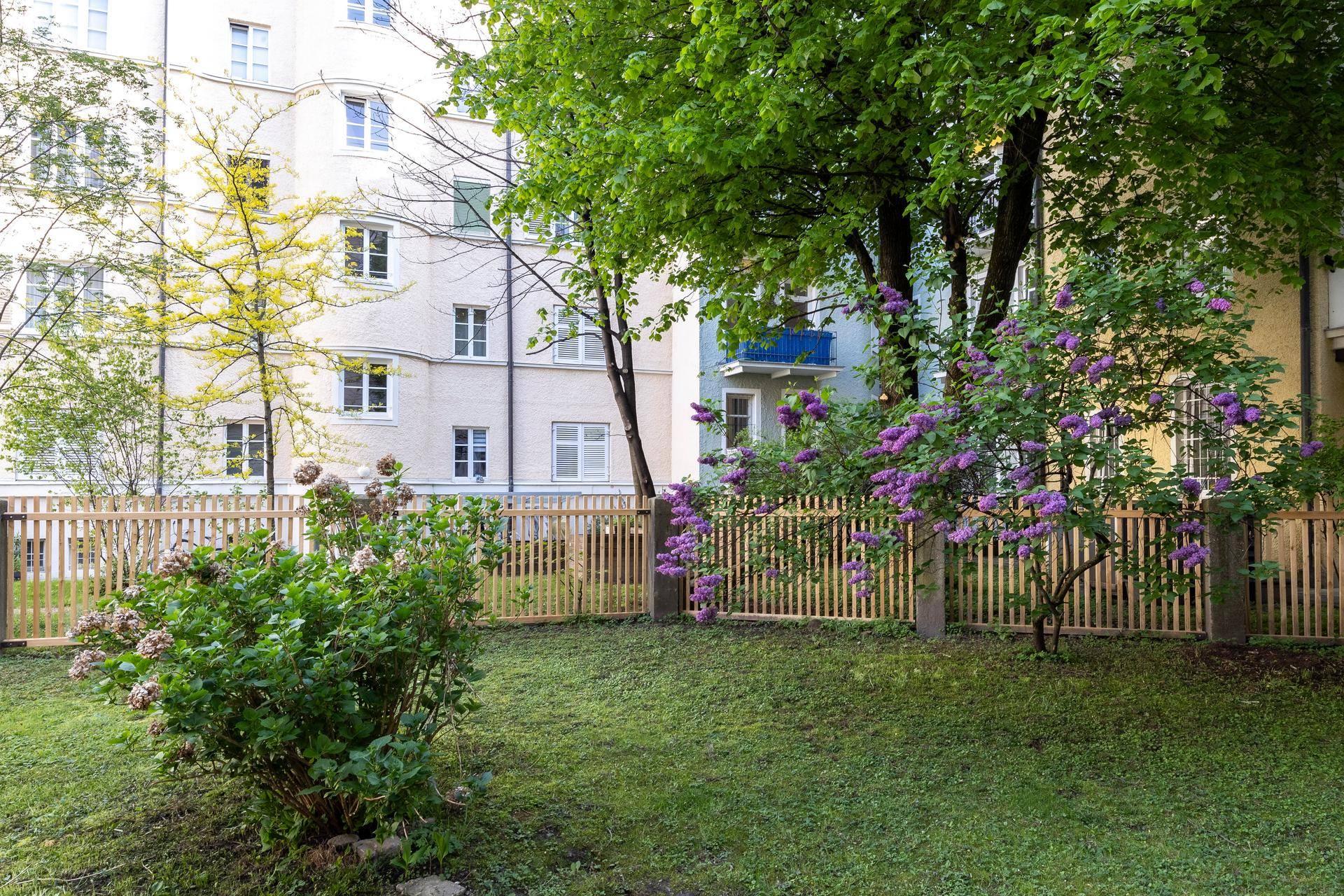 Gepflegter Gartenbereich mit Rasenfläche, Holzzaun und blühenden Sträuchern vor Wohngebäuden.