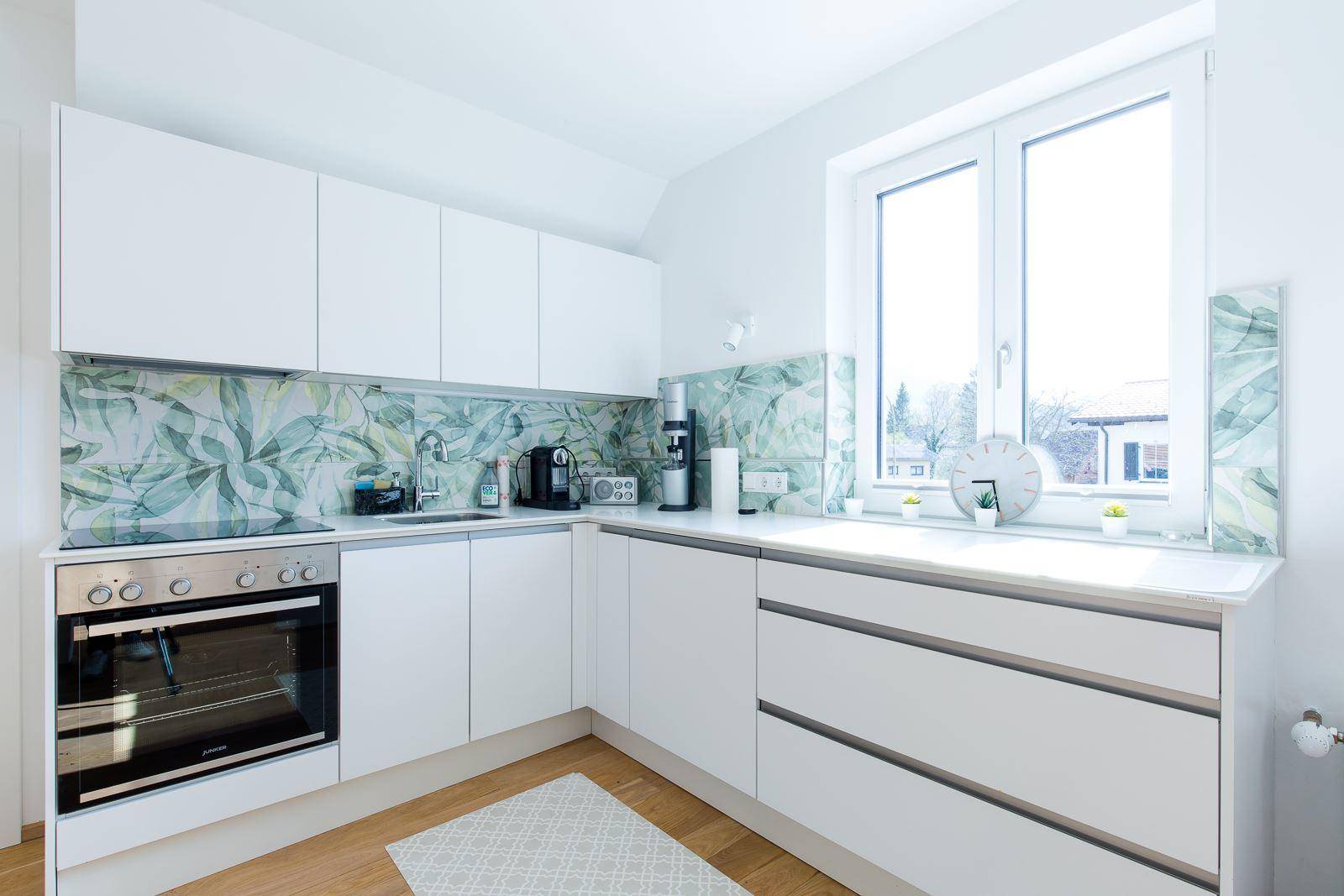 Moderne weiße Küche mit Holzboden, Fenster und floralem Fliesenspiegel.