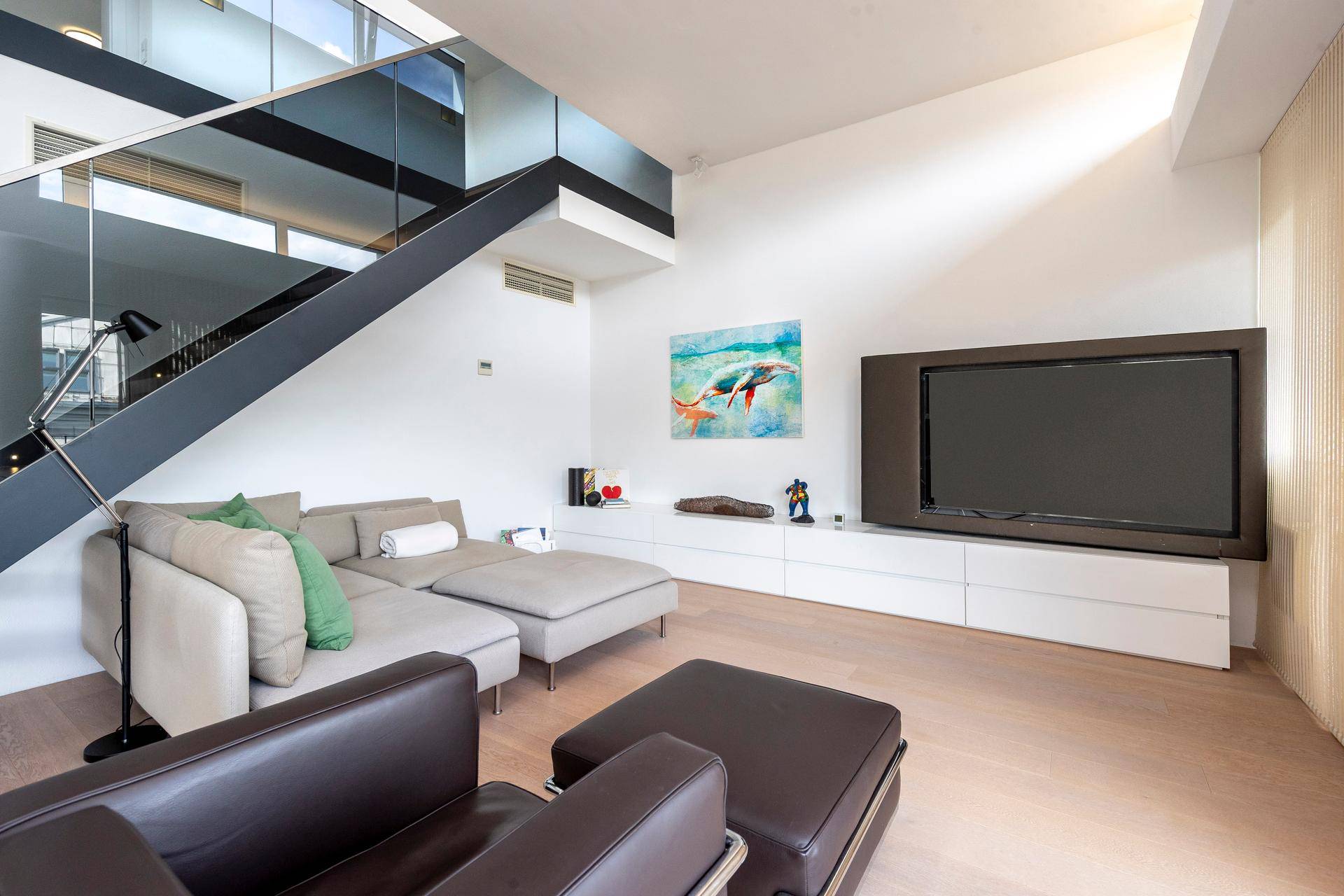 Gemütlicher Wohnbereich mit bequemer Couch, Fernseher und moderner Treppe mit Glasgeländer.