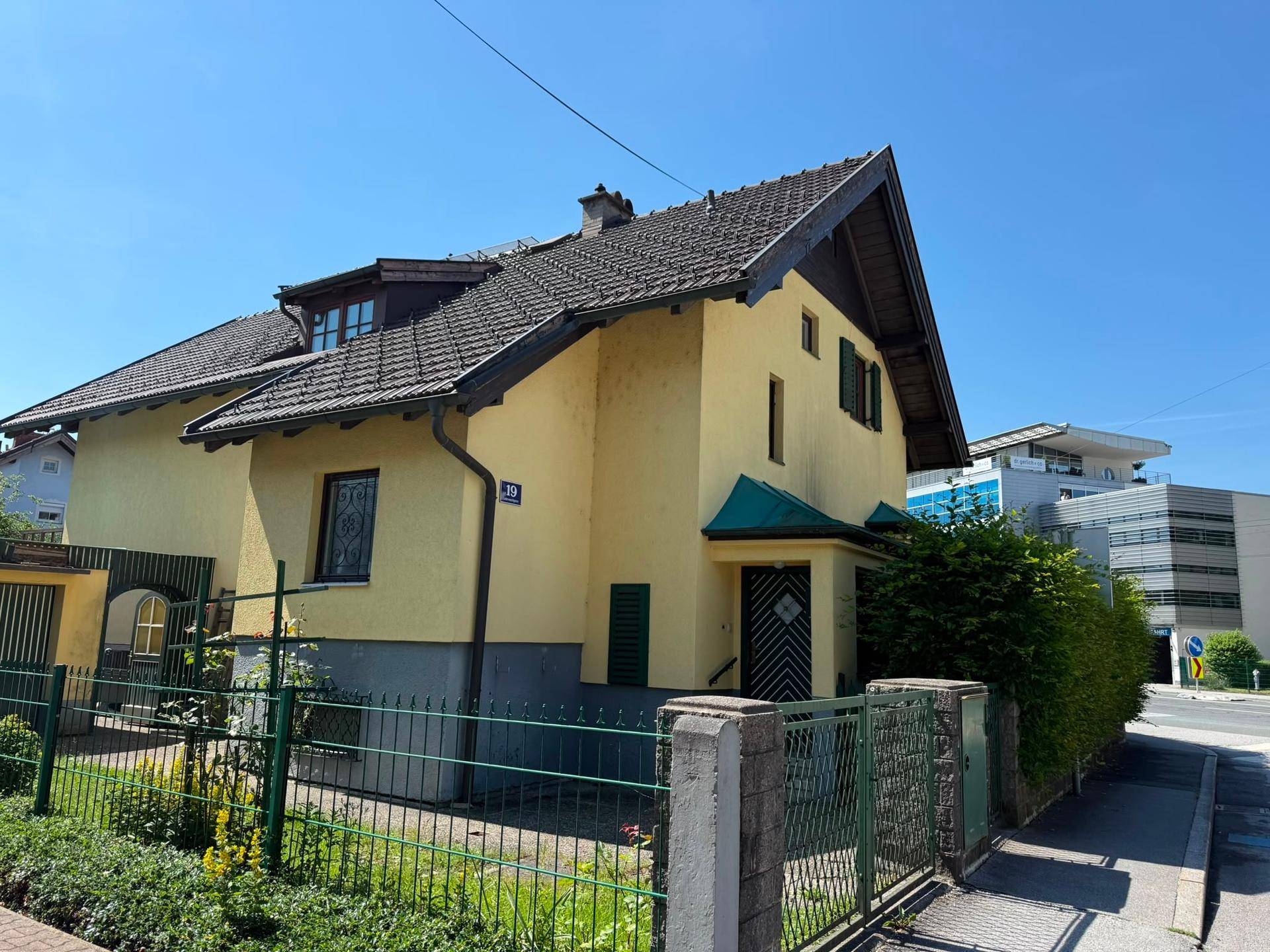 Gelbes Einfamilienhaus mit Satteldach, grünen Fensterläden und Hecke an der Straße.
