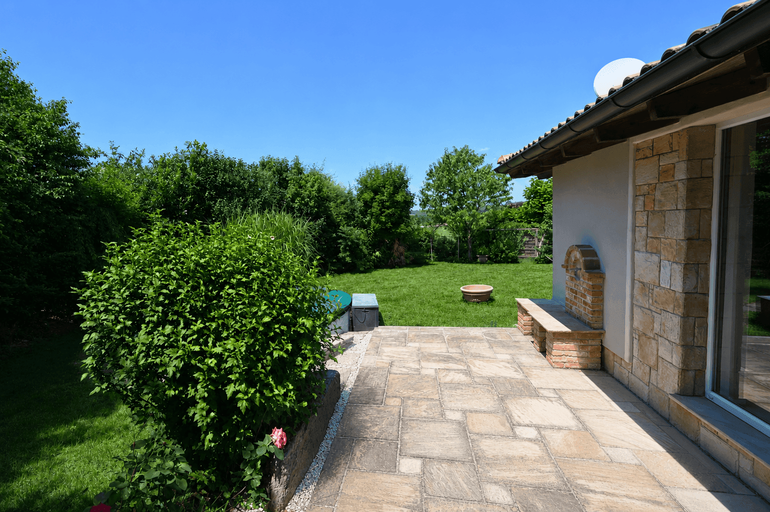 Geräumige Steinterrasse mit integriertem Grillbereich und Blick auf den gepflegten Garten.