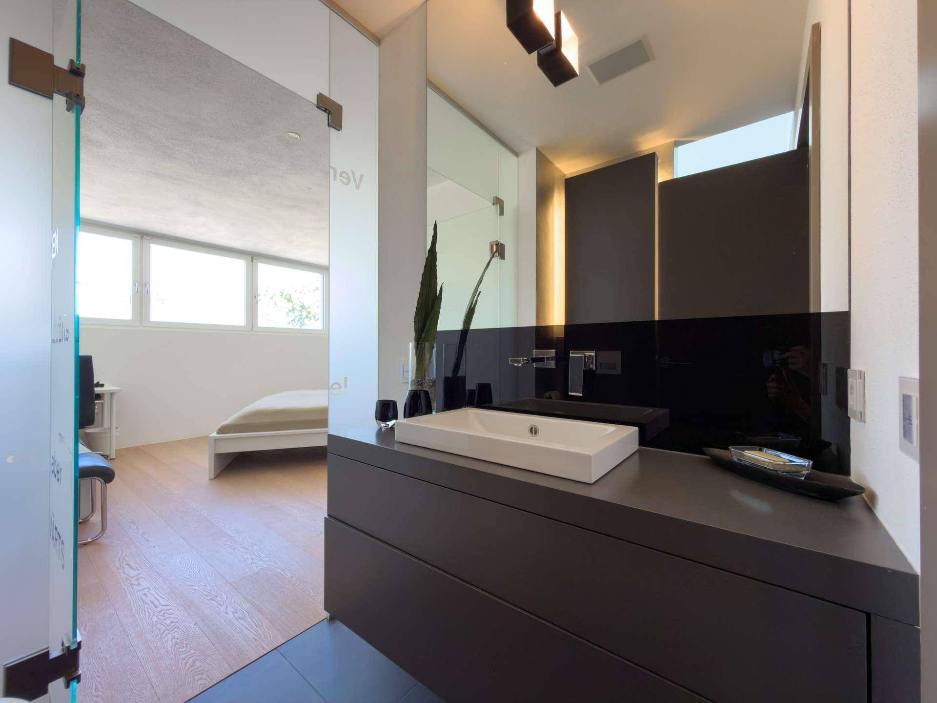 Modernes Badezimmer mit Einzelwaschbecken, dunklen Oberflächen und Blick ins Schlafzimmer.