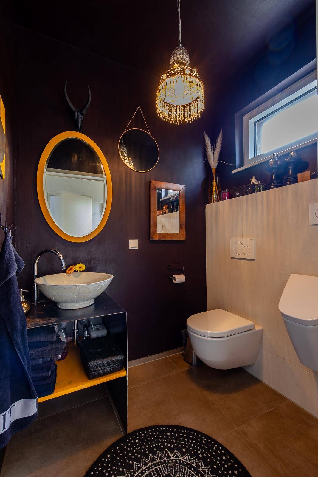 Dunkles Badezimmer mit ovalem Spiegel, Aufsatzwaschbecken und Wand-WC.