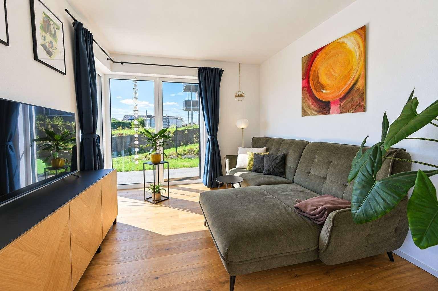 Gemütliches Wohnzimmer mit Sofa, Fernseher und großen Fenstern, die viel Licht hereinlassen.