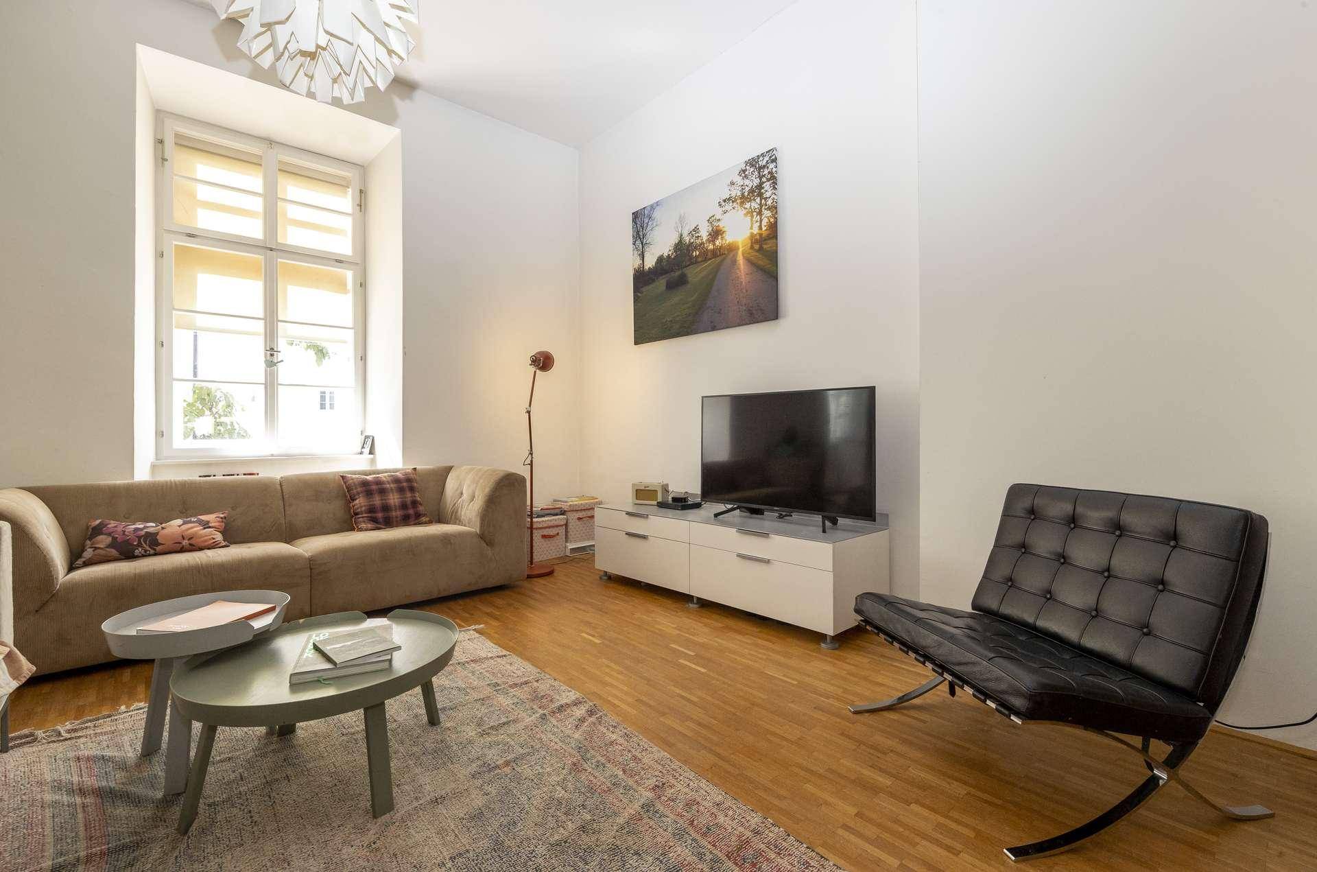 Gemütliches Wohnzimmer mit beigem Sofa, Fernseher und Holzboden, hell durch große Fenster.