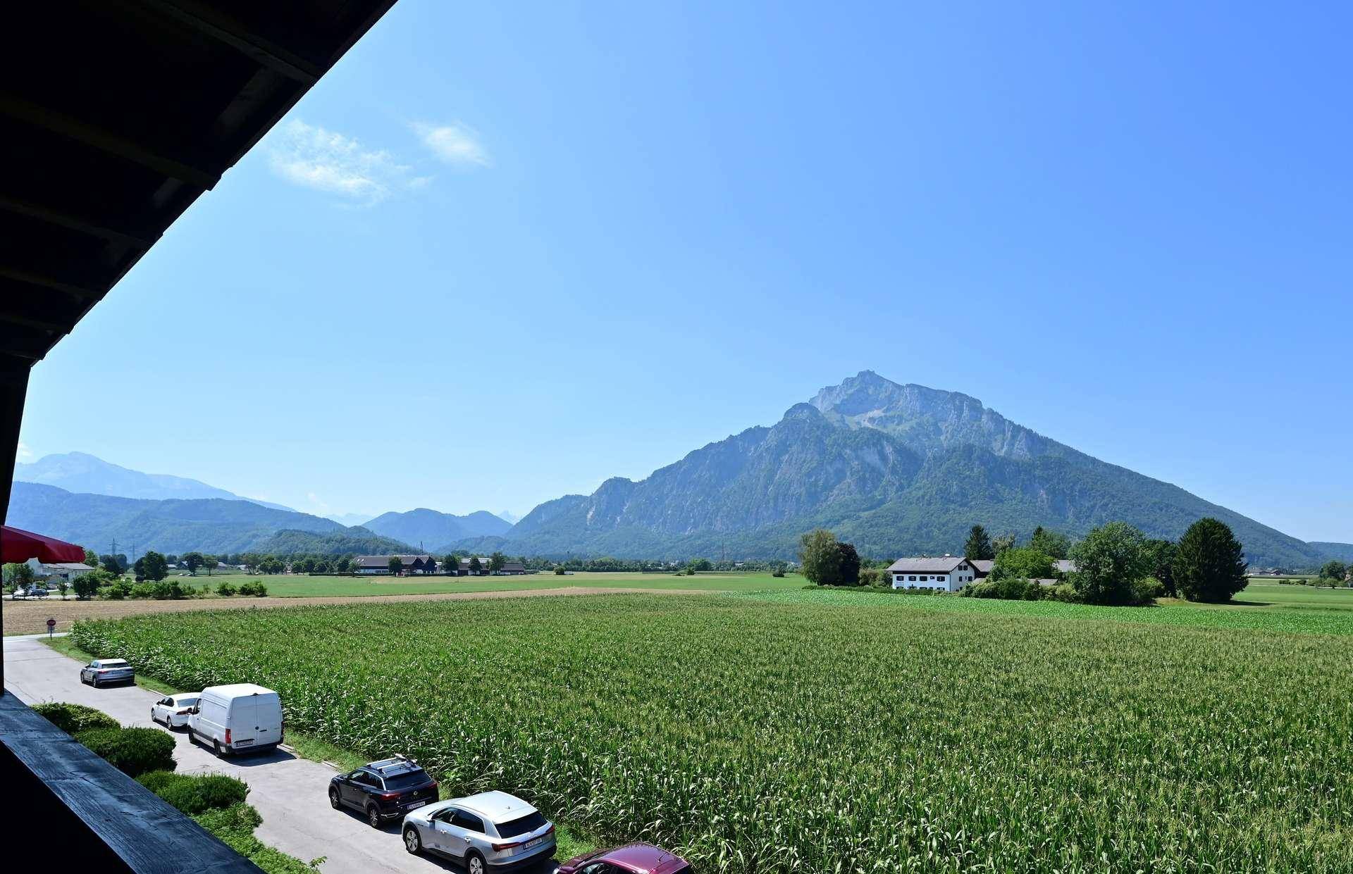 Weitblick vom Balkon über grüne Felder, Berge und eine Straße mit parkenden Autos.