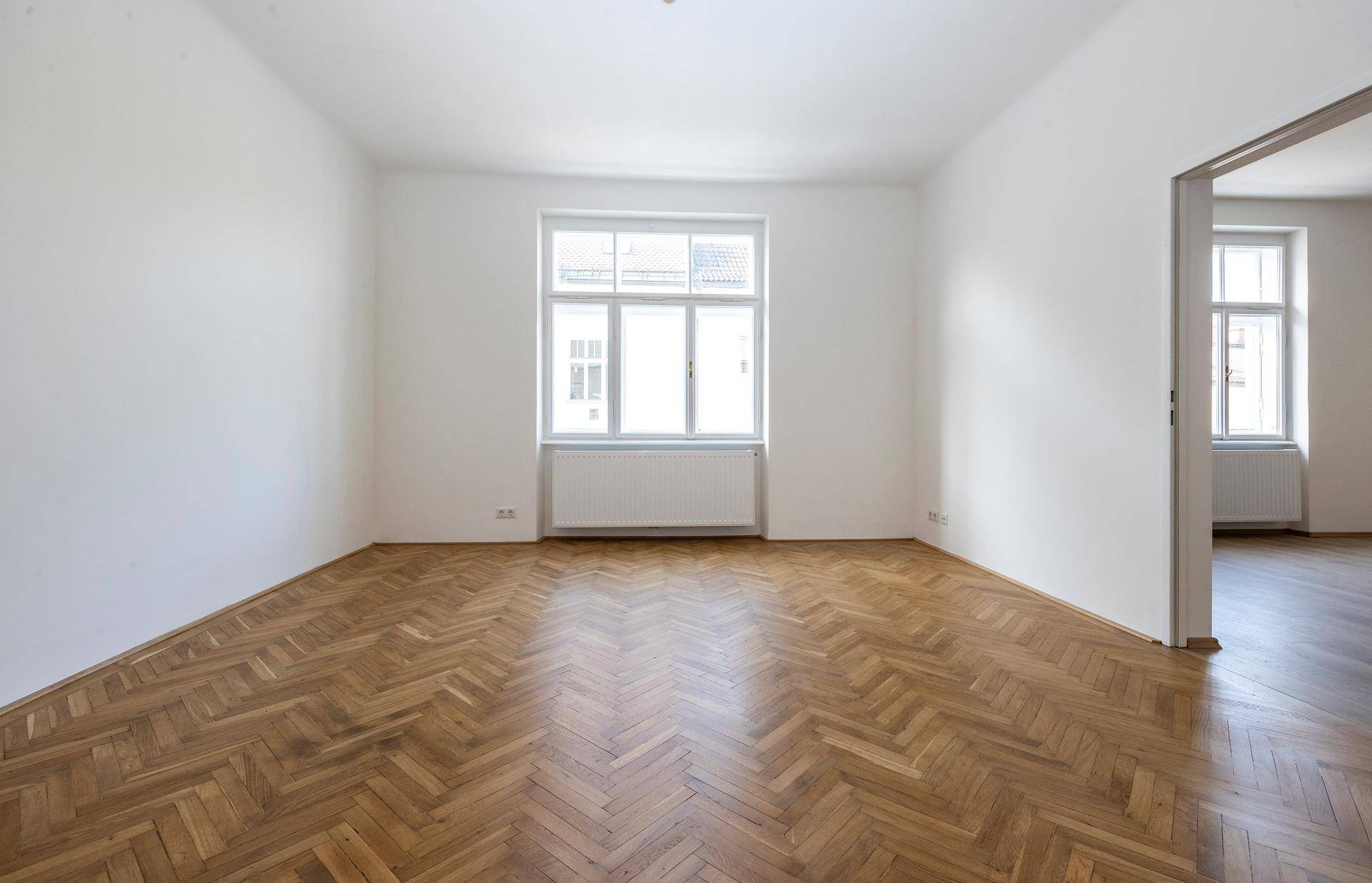 Helles, leeres Zimmer mit Fischgrätparkettboden und einem Fenster, das viel Tageslicht hereinlässt.