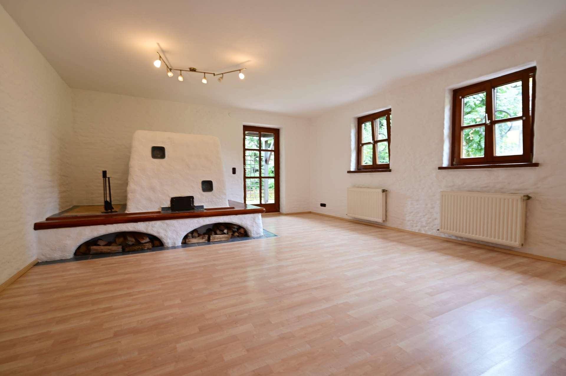 Helles Wohnzimmer mit Kachelofen, Laminatboden und zwei Fenstern mit Blick ins Grüne.