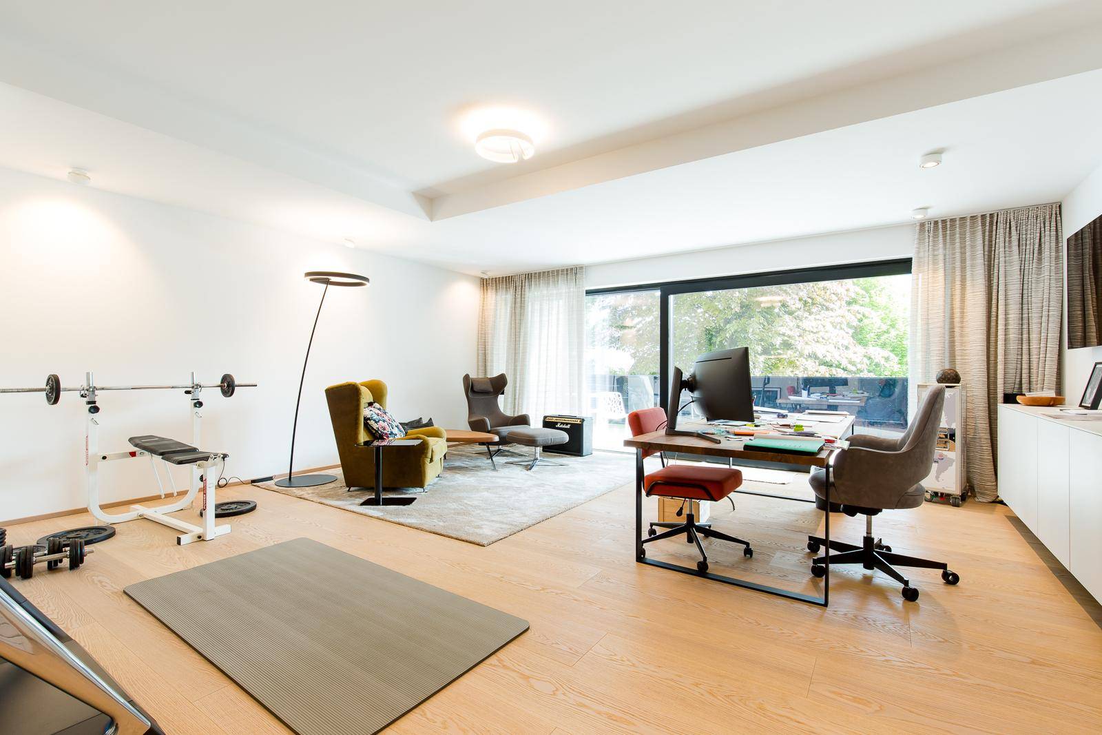 Vielseitiger Raum mit Home-Office-Bereich, Fitnessgeräten und großen Fenstern für natürliches Licht.