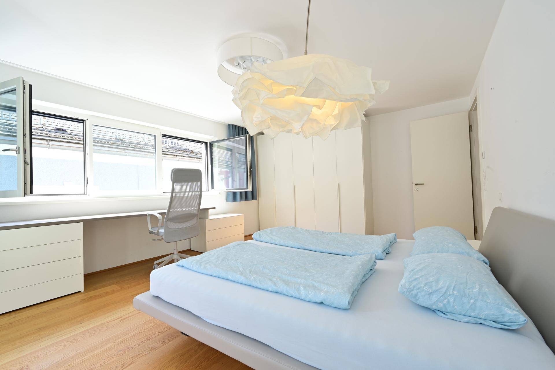 Helles Schlafzimmer mit Bett, Schreibtisch, integrierten Schränken und Fenstern mit Rollläden.