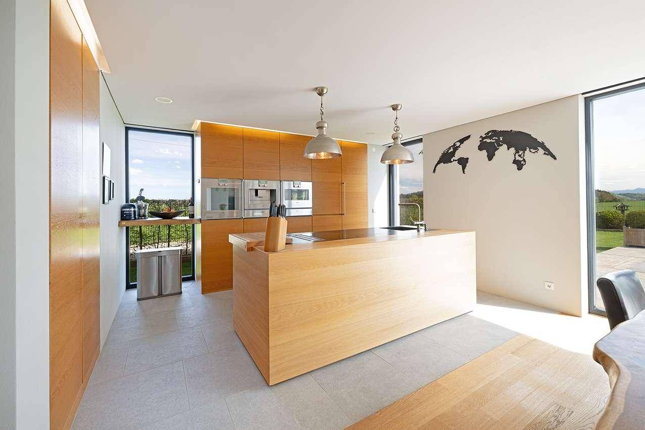 Moderne Holzküche mit Kochinsel, integrierten Geräten und viel Tageslicht durch große Fenster.