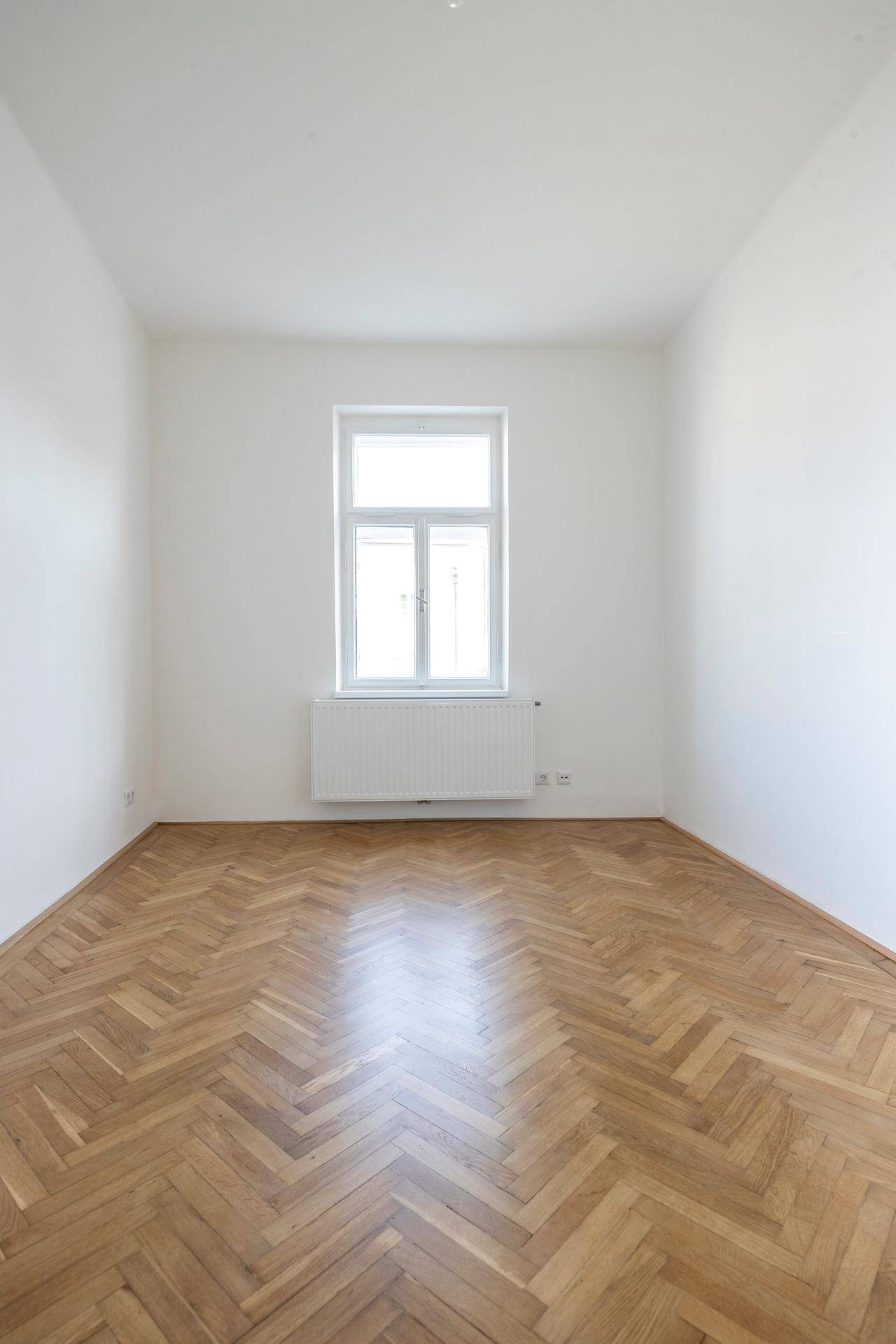 Leeres Zimmer mit elegantem Fischgrätparkett und einem Fenster, das den Raum hell erscheinen lässt.