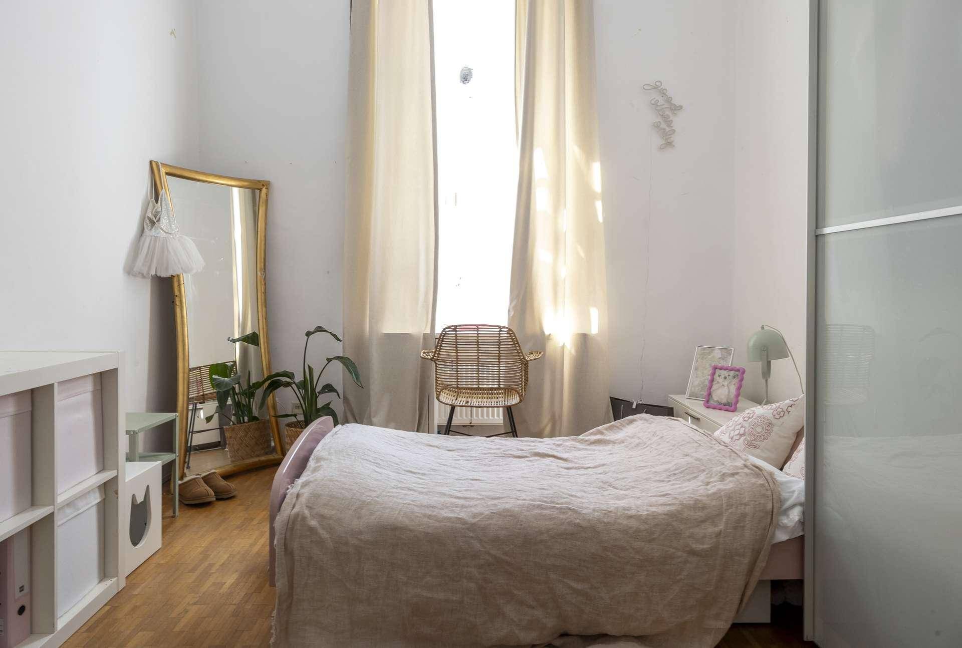 Gemütliches Schlafzimmer mit Holzboden, großem Spiegel und Vorhängen, hell durch hohes Fenster.