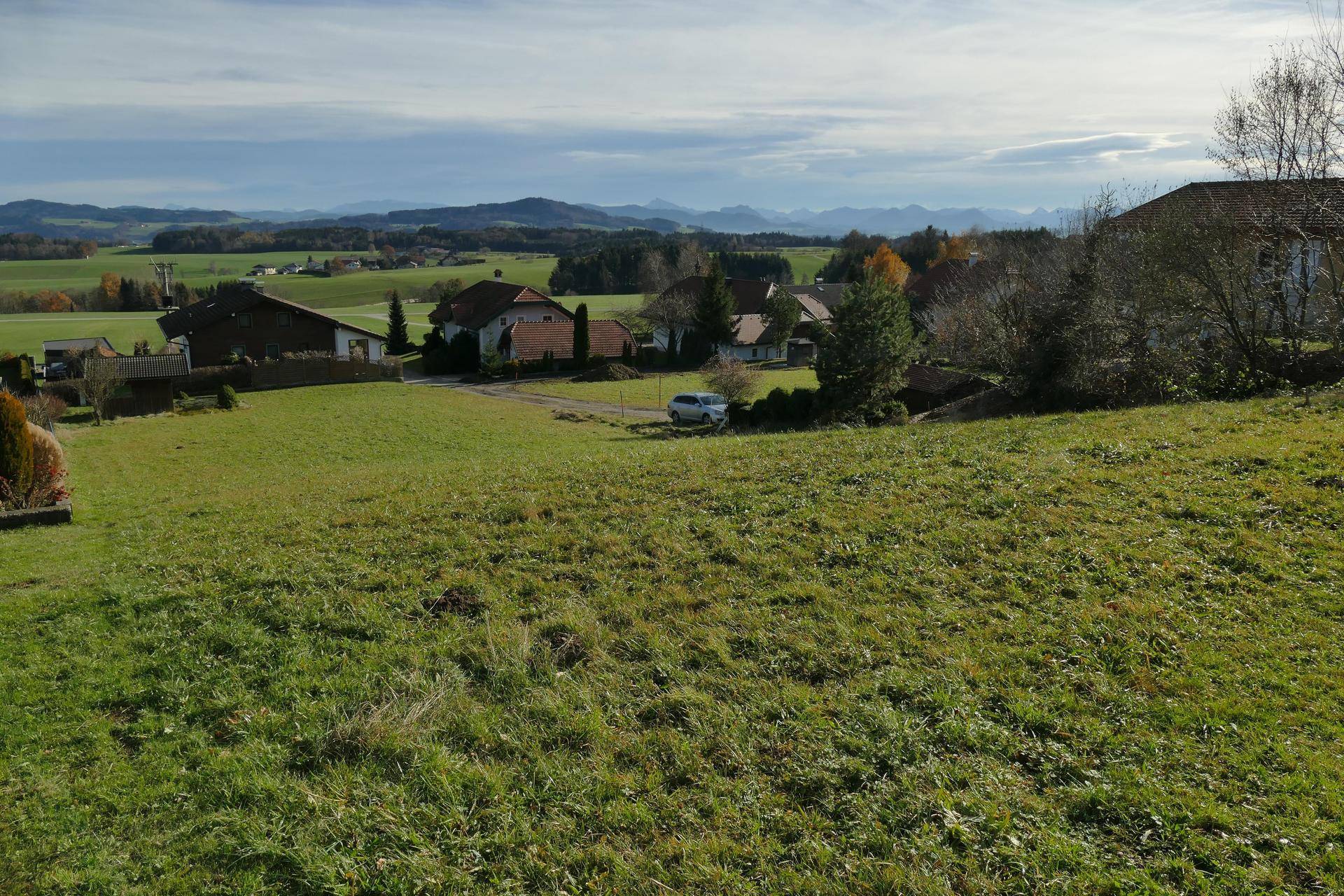 Eine weite grüne Wiese mit Häusern im Hintergrund und Bergen am Horizont unter einem bewölkten Himmel.