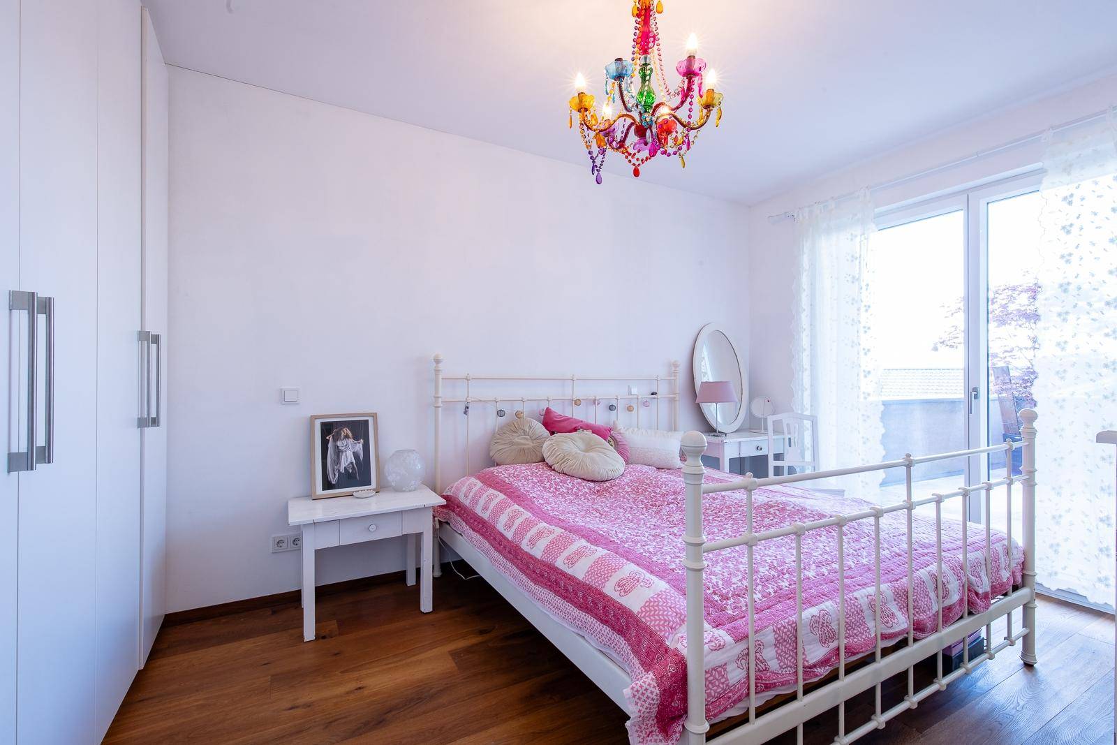 Helles Schlafzimmer mit Holzboden, weißem Bett und farbenfrohem Kronleuchter.