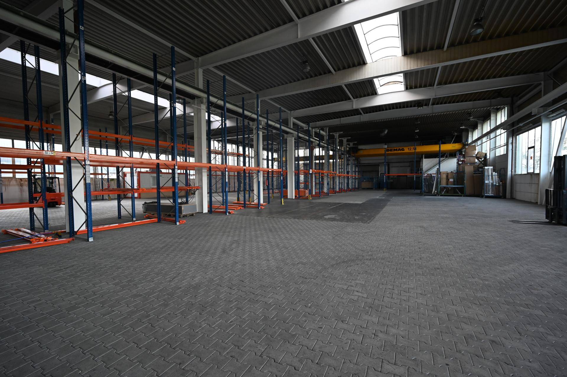 Weitläufige Lagerhalle mit leeren Regalen, Oberlichtern und einem Kran im Hintergrund.