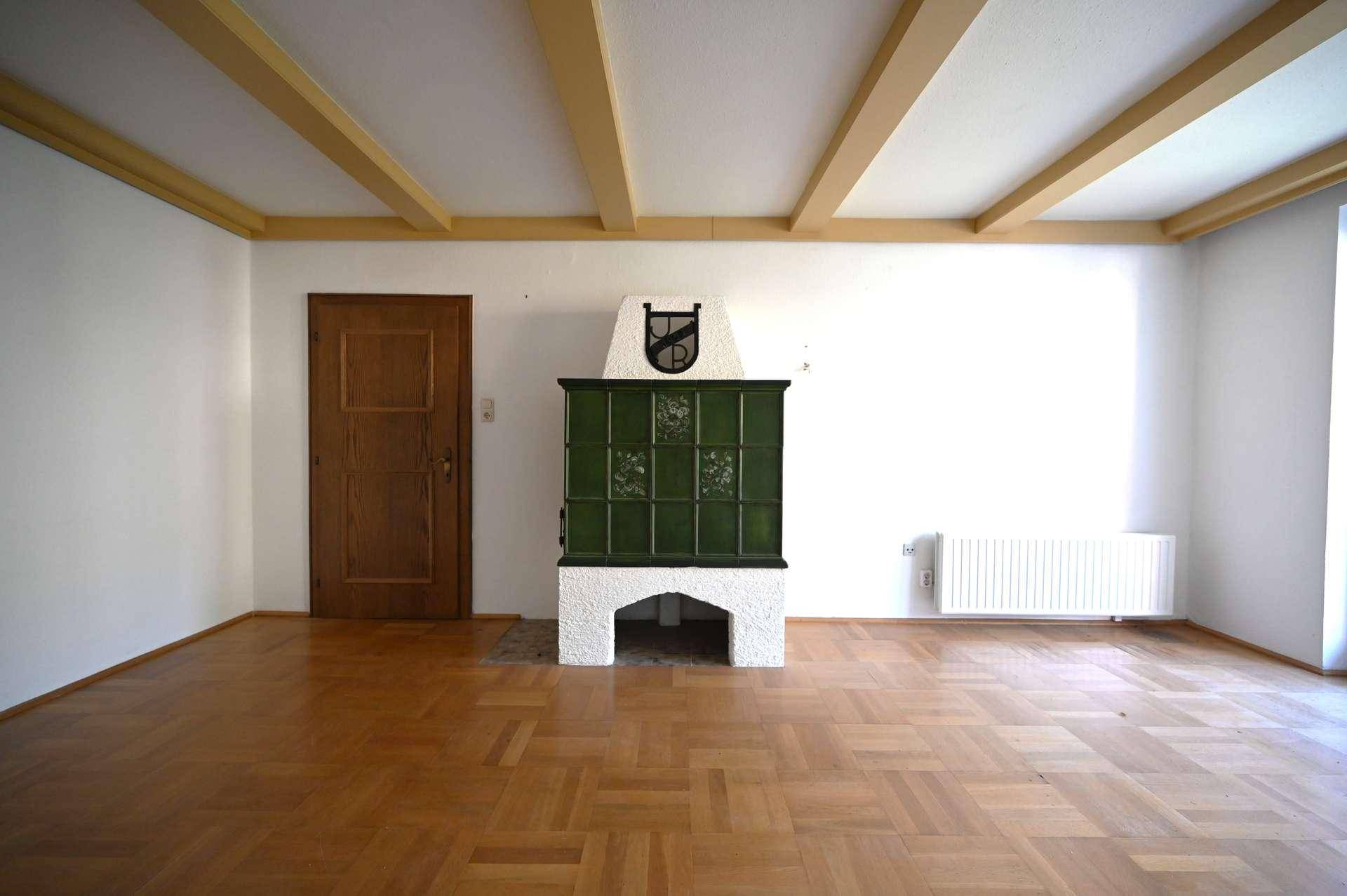 Geräumiges Wohnzimmer mit Holzboden, sichtbaren Deckenbalken und einem traditionellen Kachelofen.