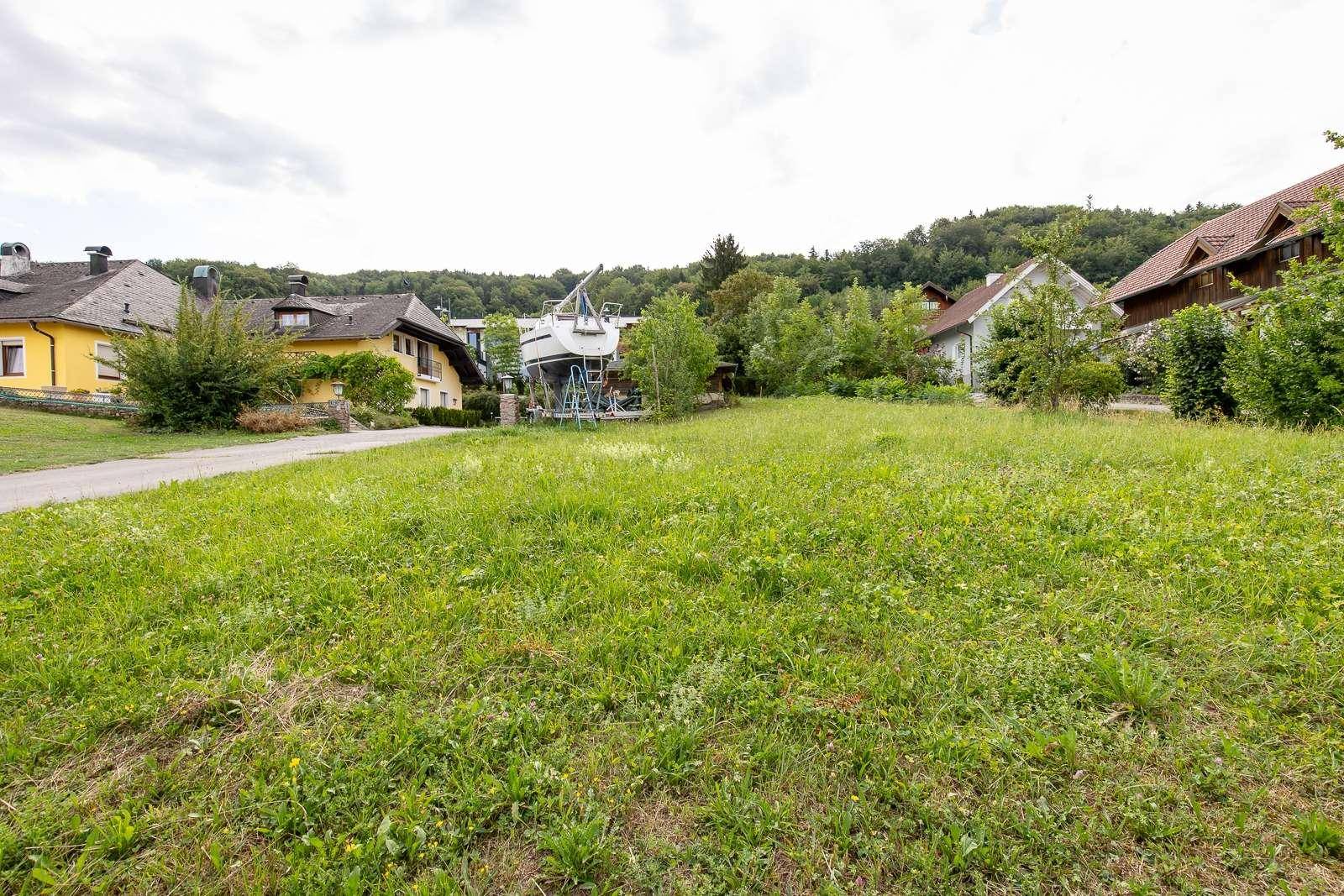 Weitläufige grüne Wiese eines Baugrundstücks mit Häusern und bewaldeten Hügeln im Hintergrund unter bewölktem Himmel.