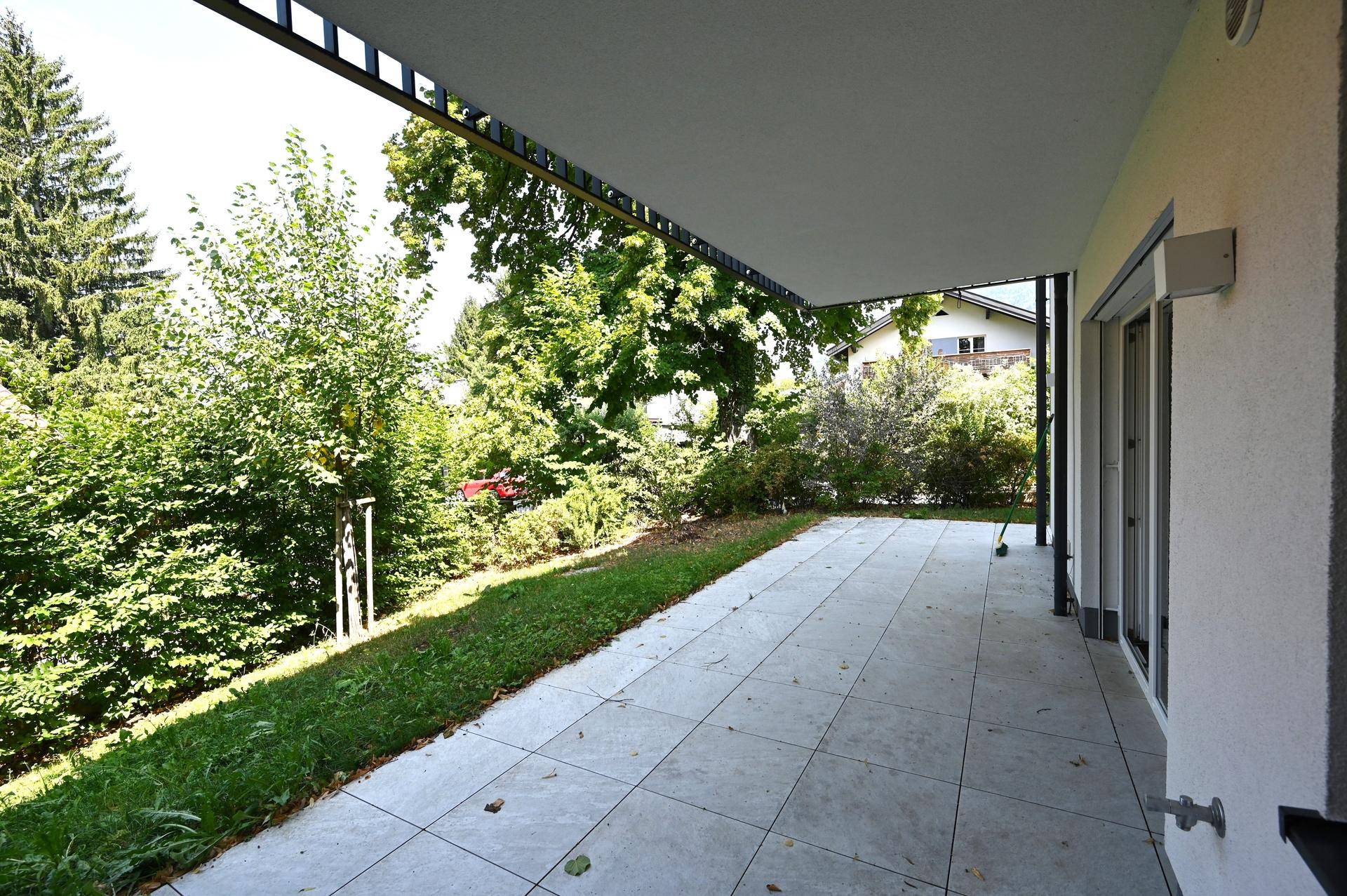 Große geflieste Terrasse mit Überdachung, umgeben von Grünflächen und Bäumen.