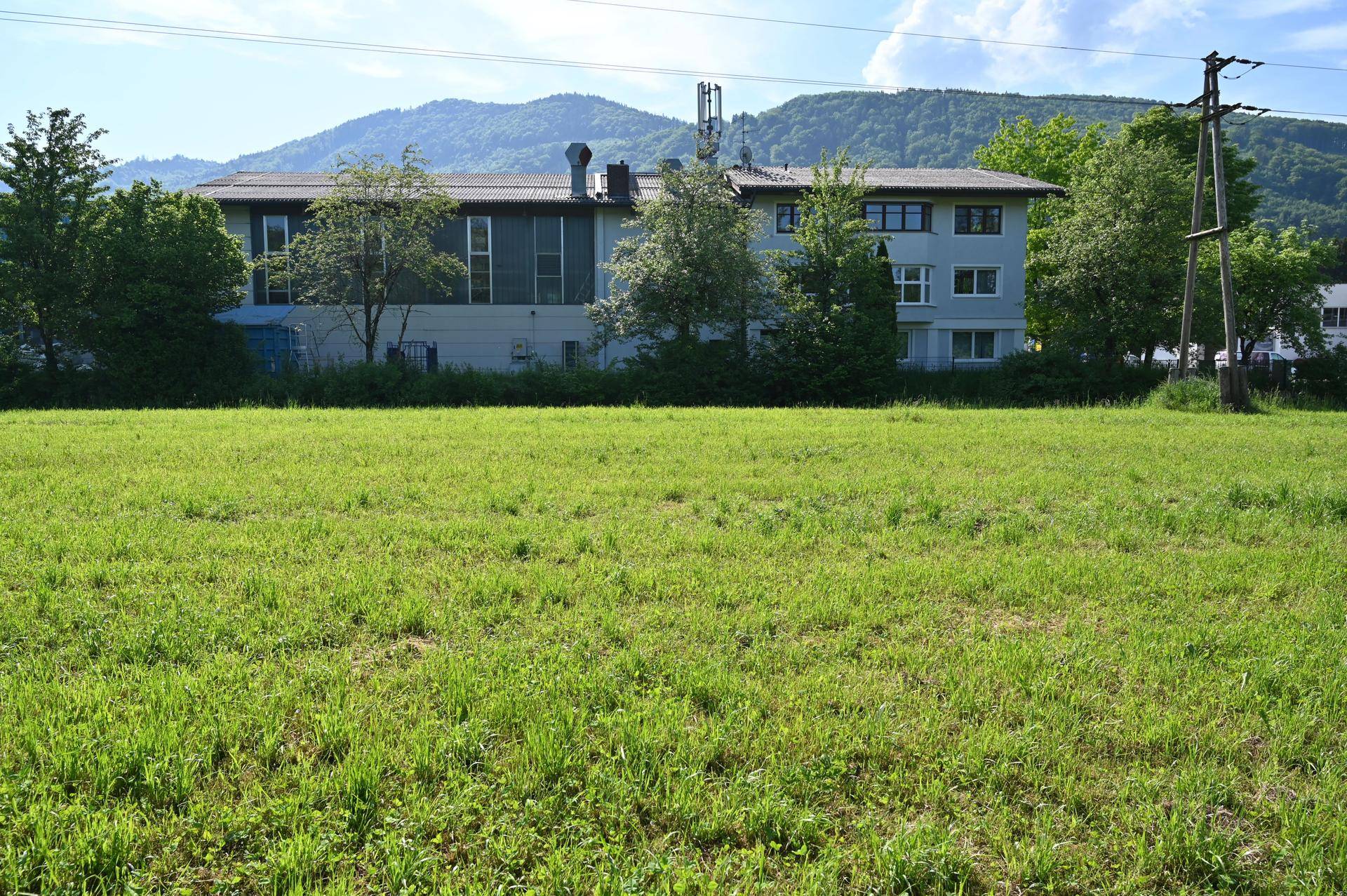 Gebäudeansicht hinter einer weiten grünen Wiese mit Bergen im Hintergrund unter blauem Himmel.