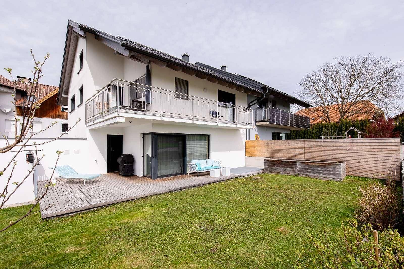 Moderne Hausfassade mit Holzterrasse, Rasenfläche und Balkonen, umgeben von Grün.