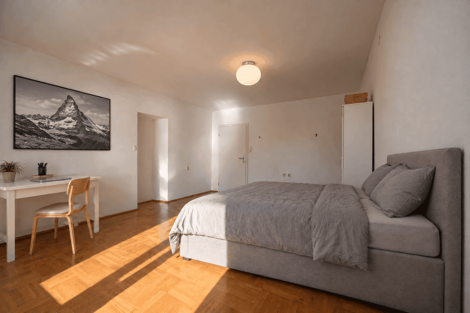Gemütliches Schlafzimmer mit Holzboden, großem Bett und Schreibtisch, durchflutet von Sonnenlicht.