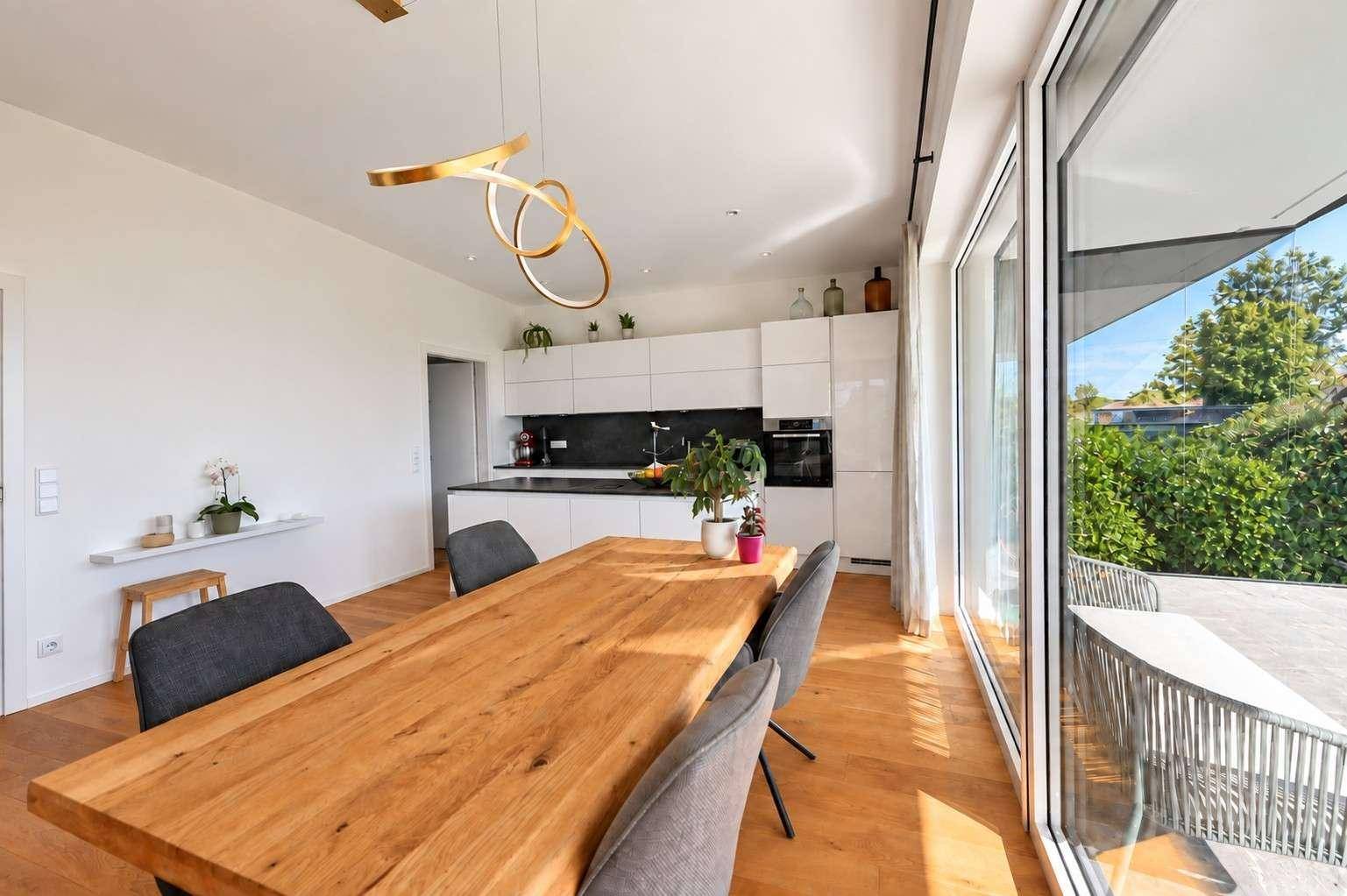 Moderne Einbauküche mit weißen Fronten und Holztisch im Essbereich, hell und offen gestaltet.