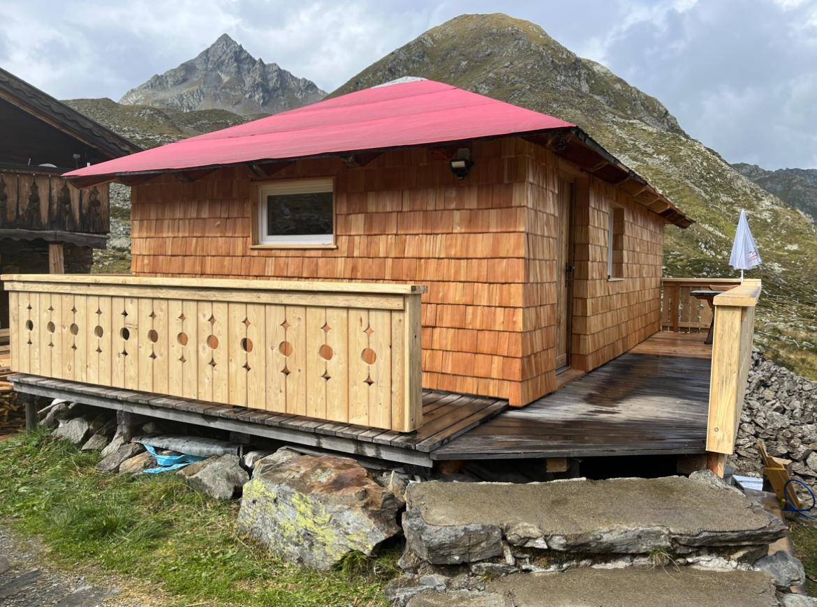 Kleine Holzhütte mit rotem Dach und einer Holzterrasse, umgeben von alpiner Landschaft und Bergen.