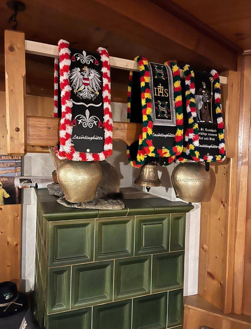 Traditioneller grüner Kachelofen mit dekorativen Kuhglocken und bestickten Bändern in einer Holzhütte.