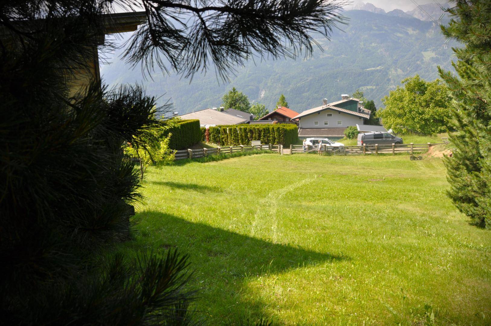 Blick über eine grüne Wiese auf benachbarte Häuser, eine Hecke und eine malerische Berglandschaft.
