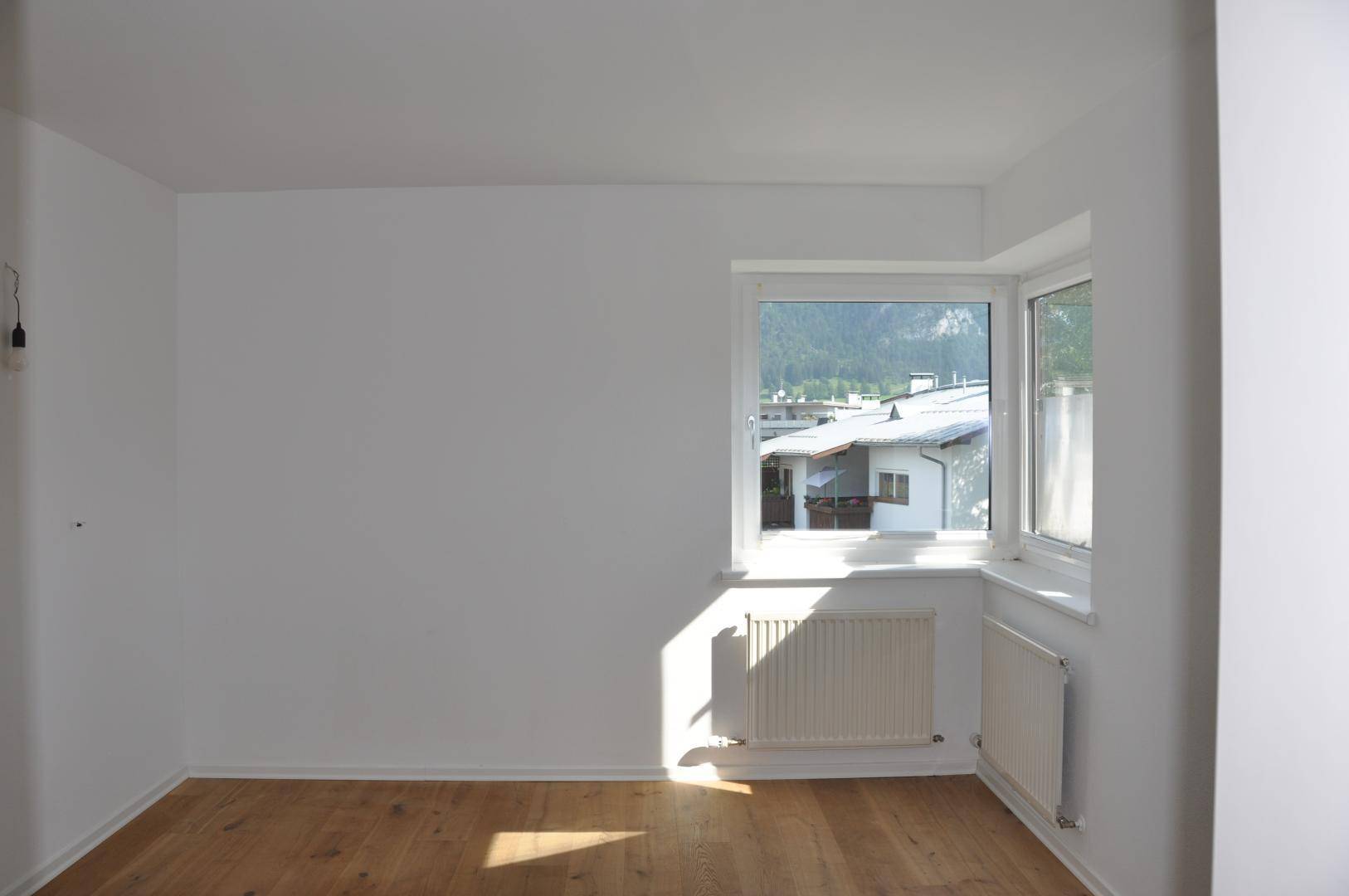 Heller, leerer Raum mit Holzboden und großen Fenstern, die viel Sonnenlicht hereinlassen.