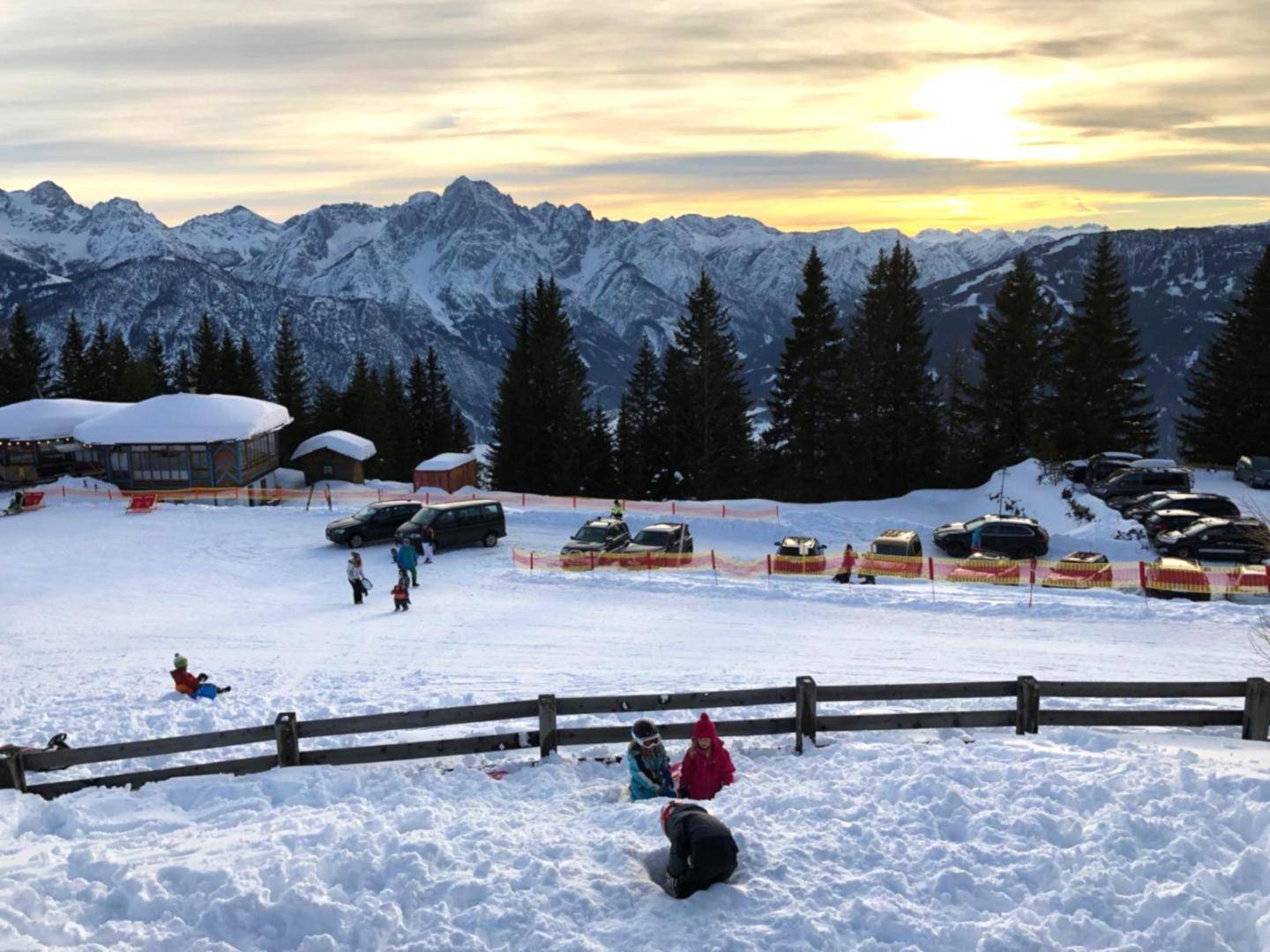Weitläufige Schneelandschaft mit spielenden Kindern, Parkplätzen und majestätischen Bergen im Hintergrund.