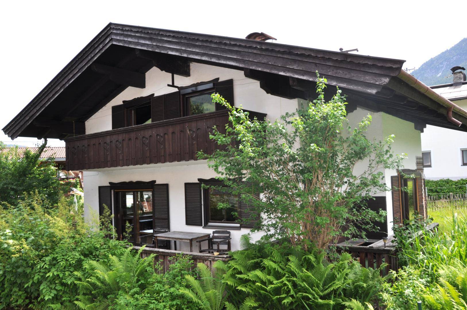 Haus mit Holzbalkon und Außensitzbereich, umgeben von üppigem Grün und Bäumen.