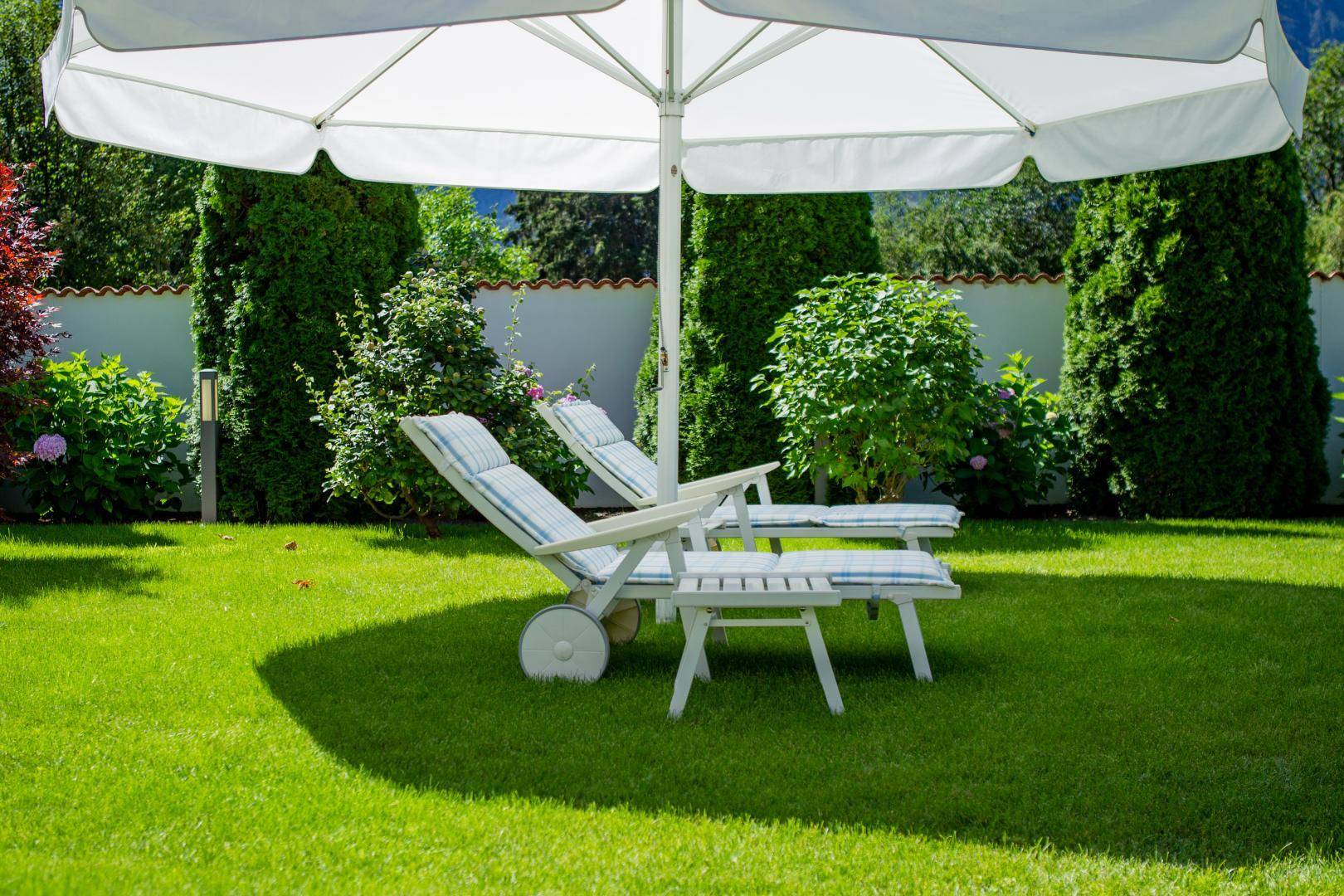 Gemütliche Sonnenliegen unter einem großen Sonnenschirm auf der grünen Rasenfläche im Garten.