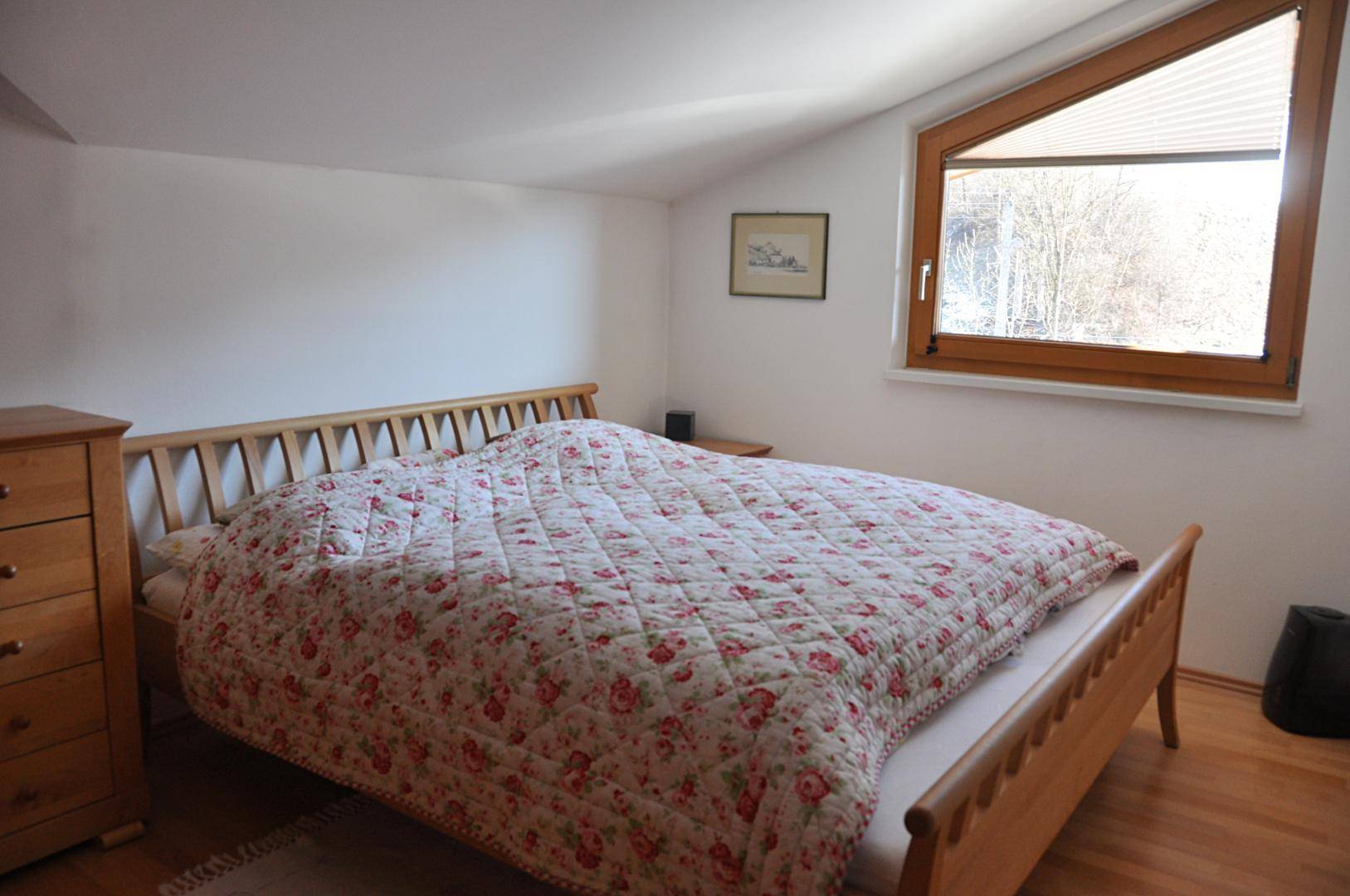 Gemütliches Schlafzimmer mit Holzbett, floraler Bettwäsche und einem großen Schrägfenster.