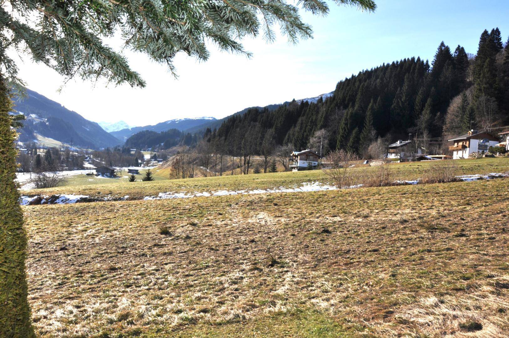Idyllische Landschaft mit grünen Wiesen, vereinzelten Schneeflecken und traditionellen Häusern vor Bergkulisse.