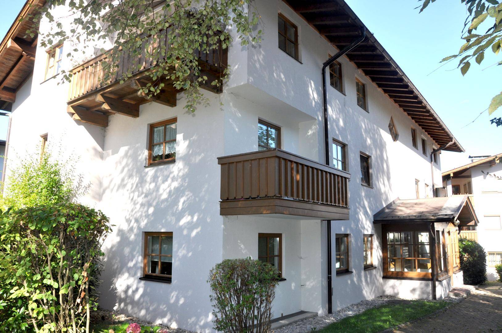 Weißes Gebäude mit traditionellen Holzbalkonen und grüner Bepflanzung im Vordergrund.