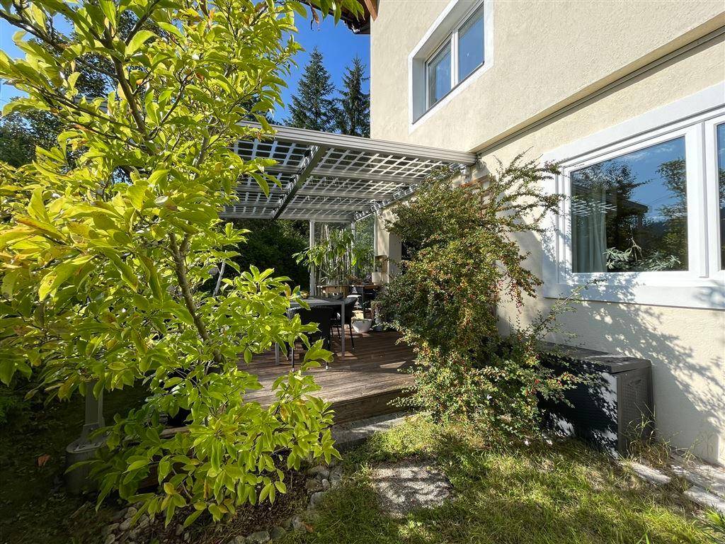 Hausfassade mit überdachter Terrasse und üppiger Bepflanzung im Gartenbereich.