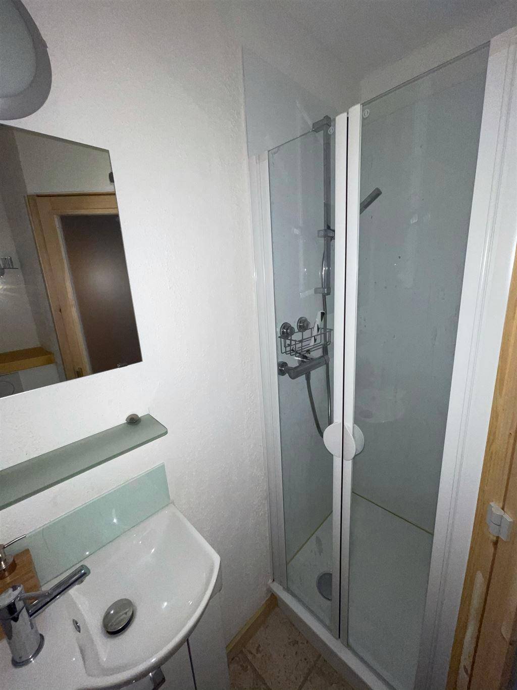 Kleines Badezimmer mit Waschbecken und einer Duschkabine, hell gefliest.
