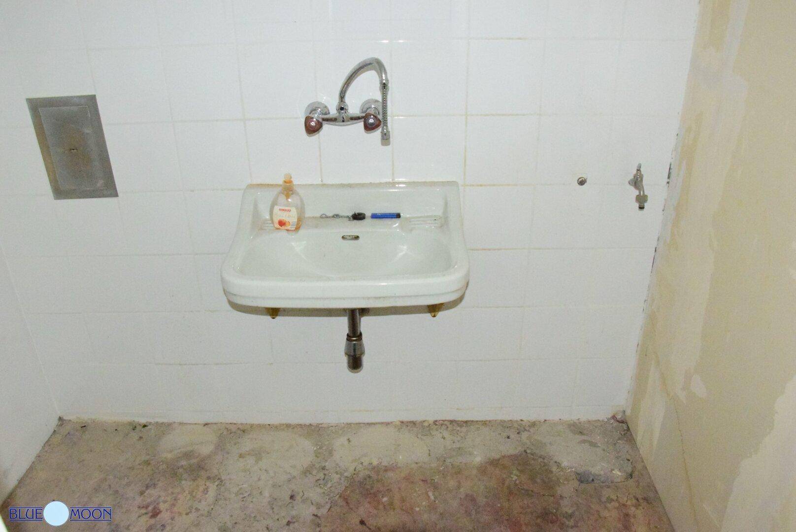 Einfaches Waschbecken an einer gefliesten Wand in einem Raum, der Renovierung benötigt, mit sichtbaren Wasseranschlüssen.