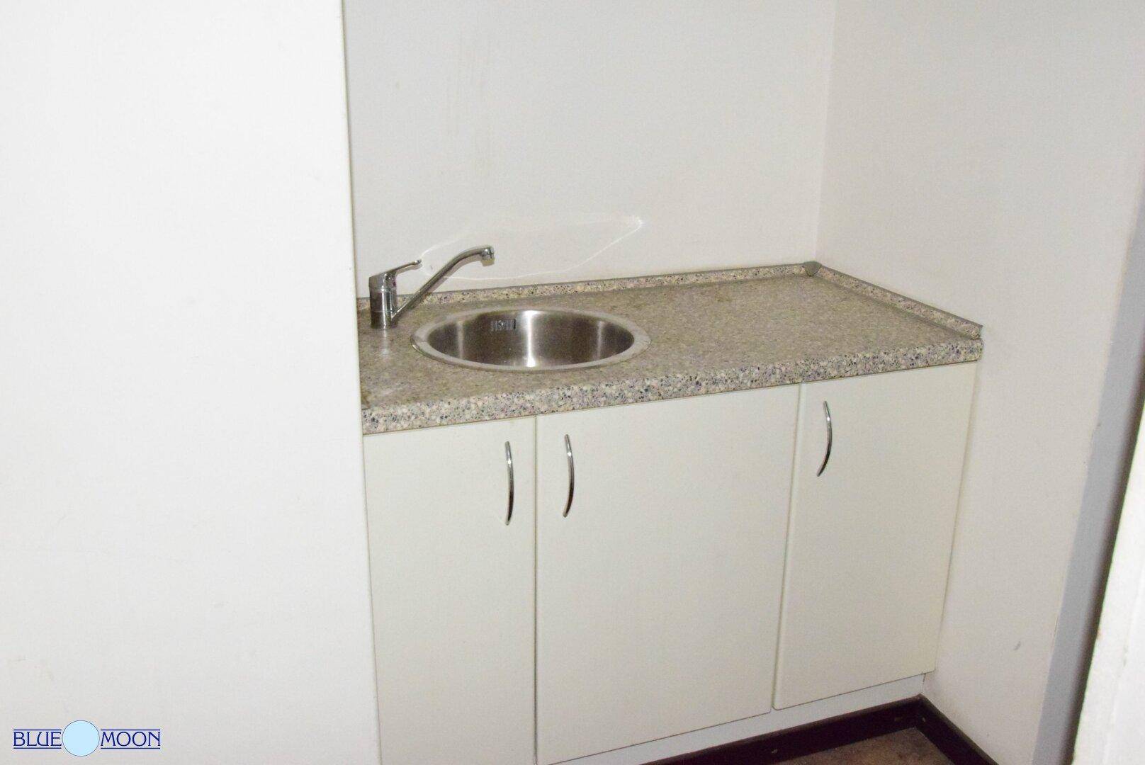 Kleine Küchenzeile mit Edelstahlspüle, Granit-Arbeitsplatte und weißen Unterschränken in einem schlichten Raum.