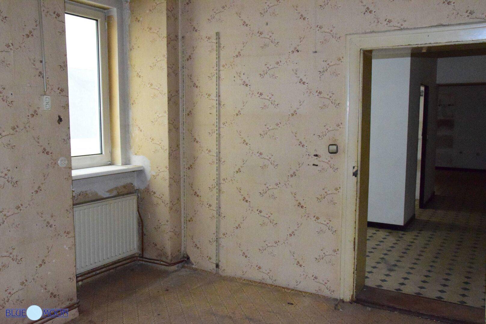 Raum mit gemusterter Tapete, einem Fenster und einem Heizkörper, der auf Renovierungsbedarf hindeutet.