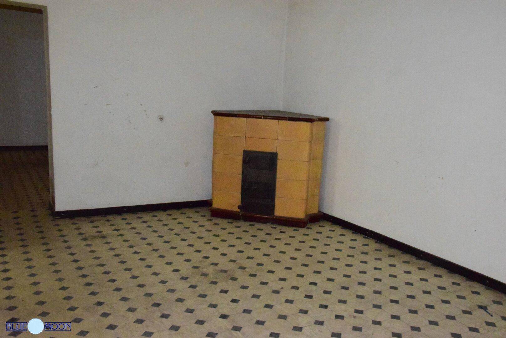 Leerer Raum mit gemustertem Fliesenboden, weißen Wänden und einem traditionellen Kachelofen in der Ecke.