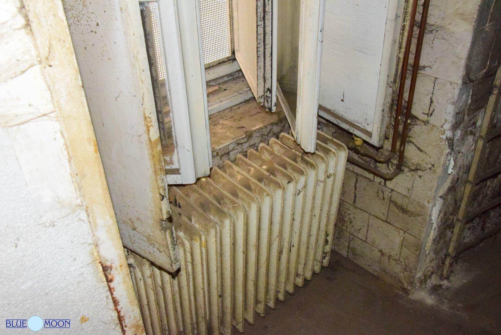 Alter Heizkörper unter einem Fenster in einem Raum mit abgenutzten Wänden, der auf Renovierungsbedarf hindeutet.
