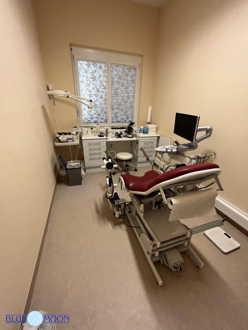Medizinischer Untersuchungsraum mit Spezialstuhl, Geräten und einem Fenster mit Jalousien.